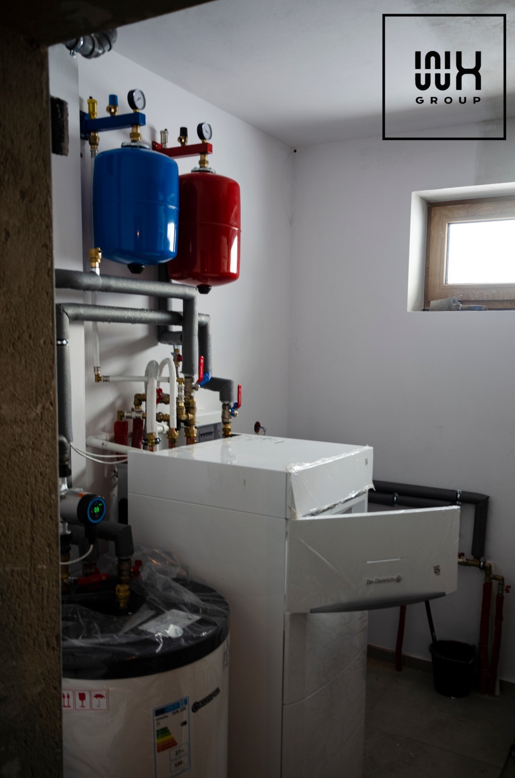 Nowoczesna instalacja grzewcza w kotłowni: biały piec, zbiornik wody, rury, manometry. Elementy jeszcze w folii ochronnej. Okno w tle.