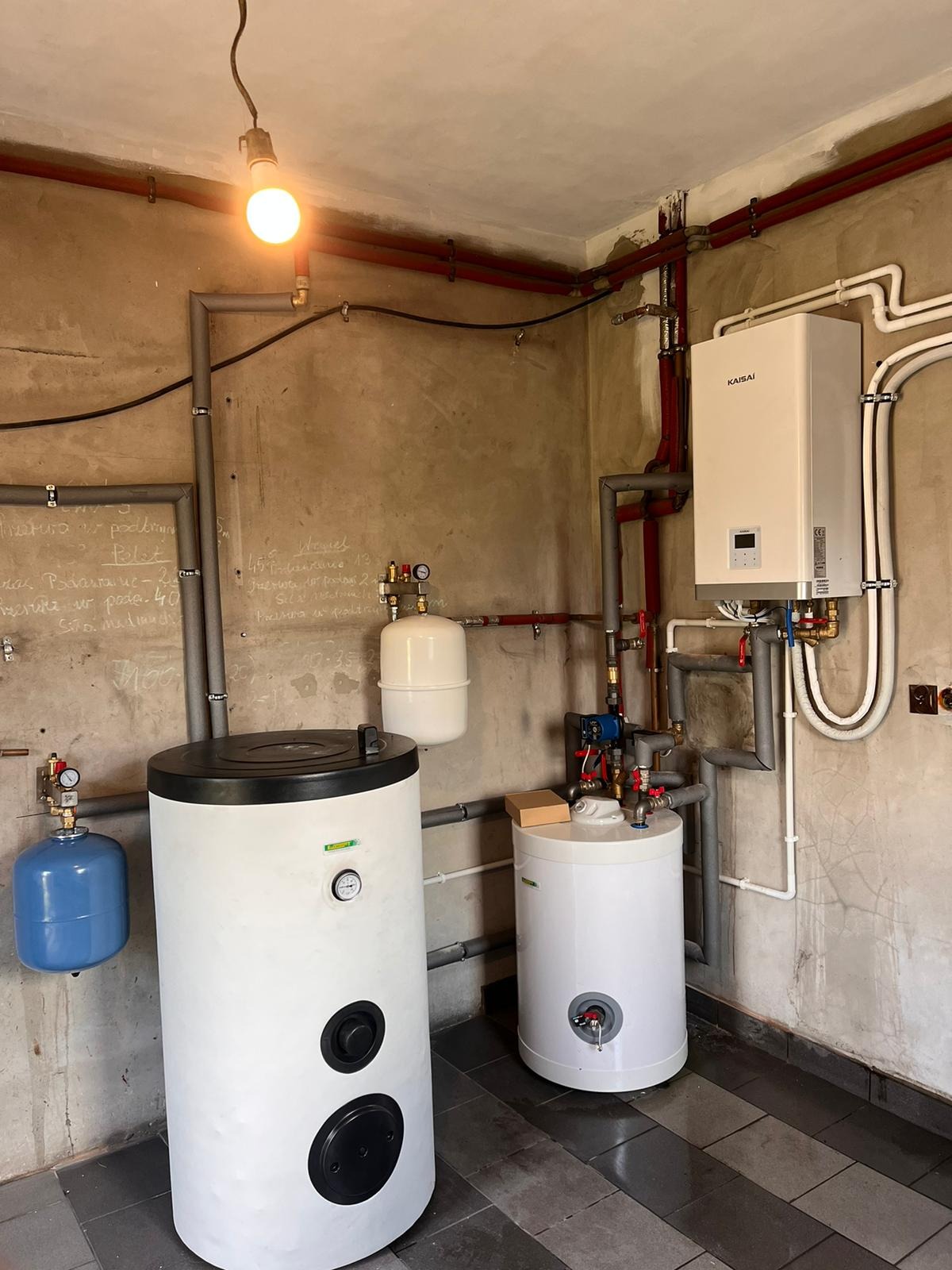 Kompletna instalacja grzewcza w kotłowni: biały piec Kaisai, zbiorniki buforowe, rury w kolorze czerwonym i szarym. Instalacja w surowym wnętrzu z betonowymi ścianami.
