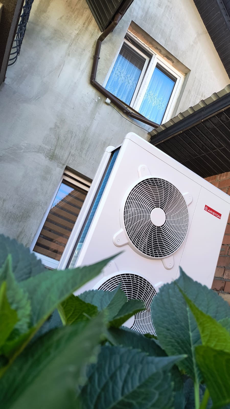 Biała, dwuczęściowa pompa ciepła Ariston zamontowana na elewacji budynku, widoczna zza zielonych liści. Ujęcie z dołu, na tle okien i fragmentu dachu.