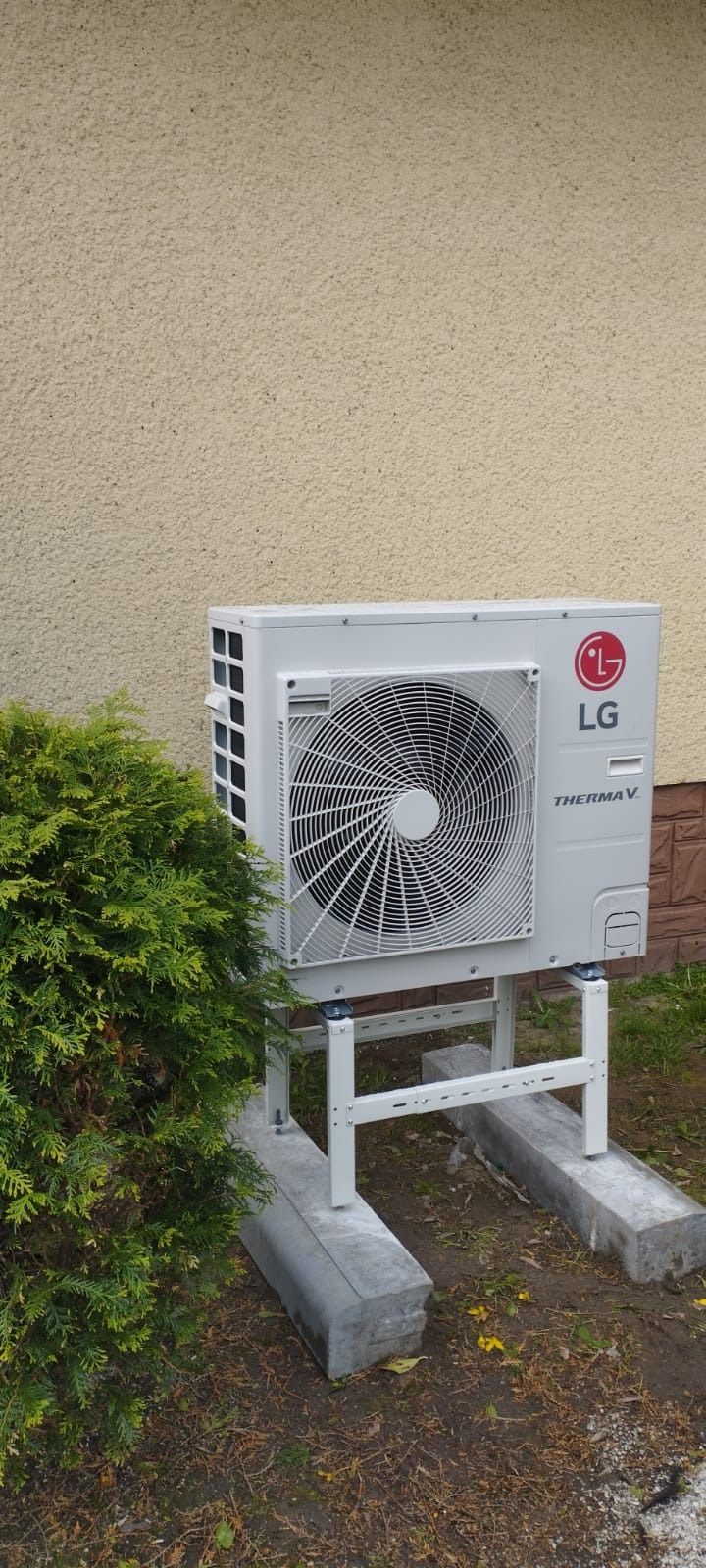 Biała zewnętrzna jednostka pompy ciepła LG Therma V, zamontowana na metalowym stelażu, z krzewem z lewej strony i beżową ścianą w tle.