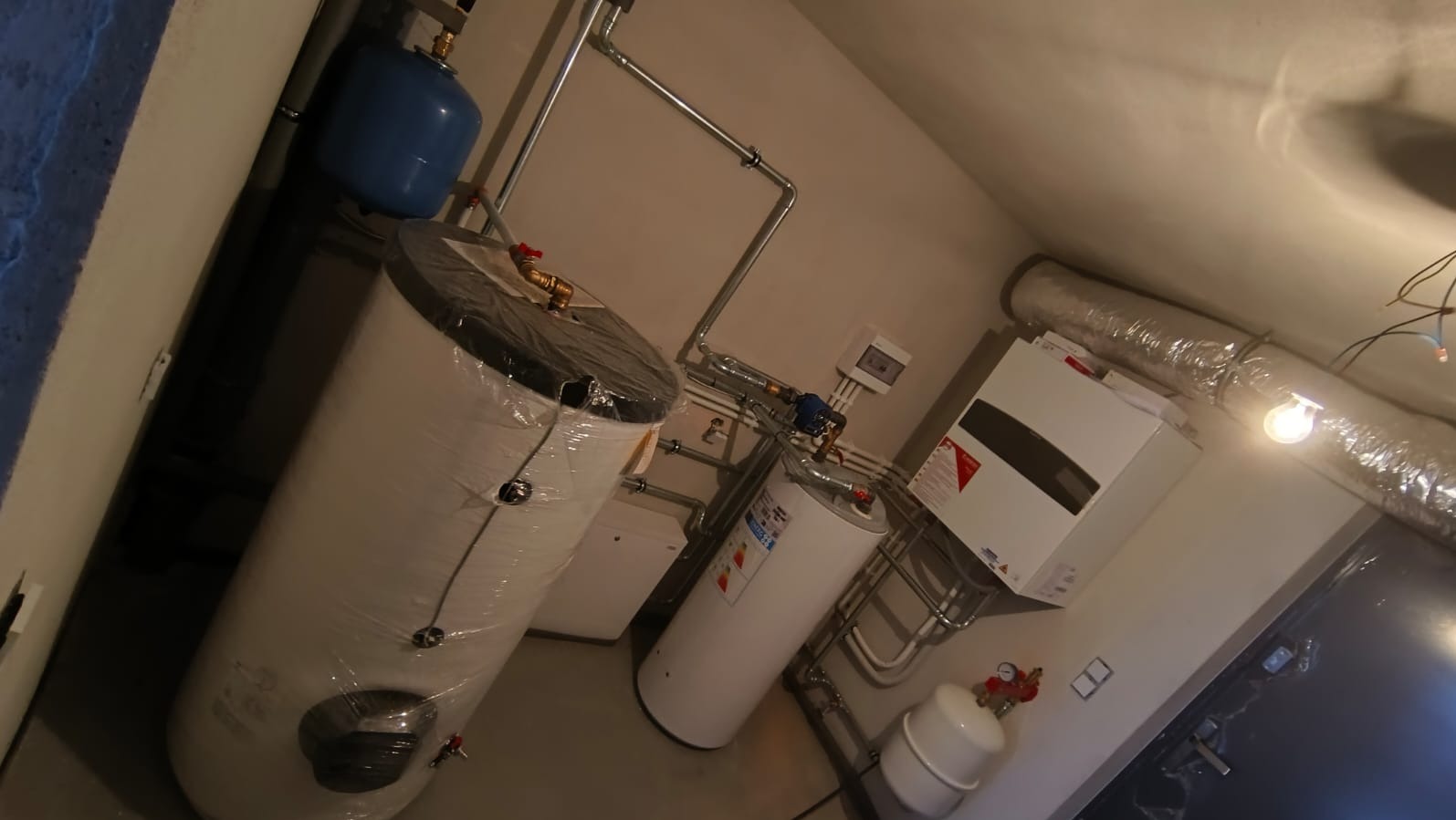 Zmontowana instalacja grzewcza w kotłowni: dwa białe zbiorniki CWU, niebieski zbiornik przeponowy i piec gazowy z podłączeniami rur. Jeden zbiornik owinięty folią.