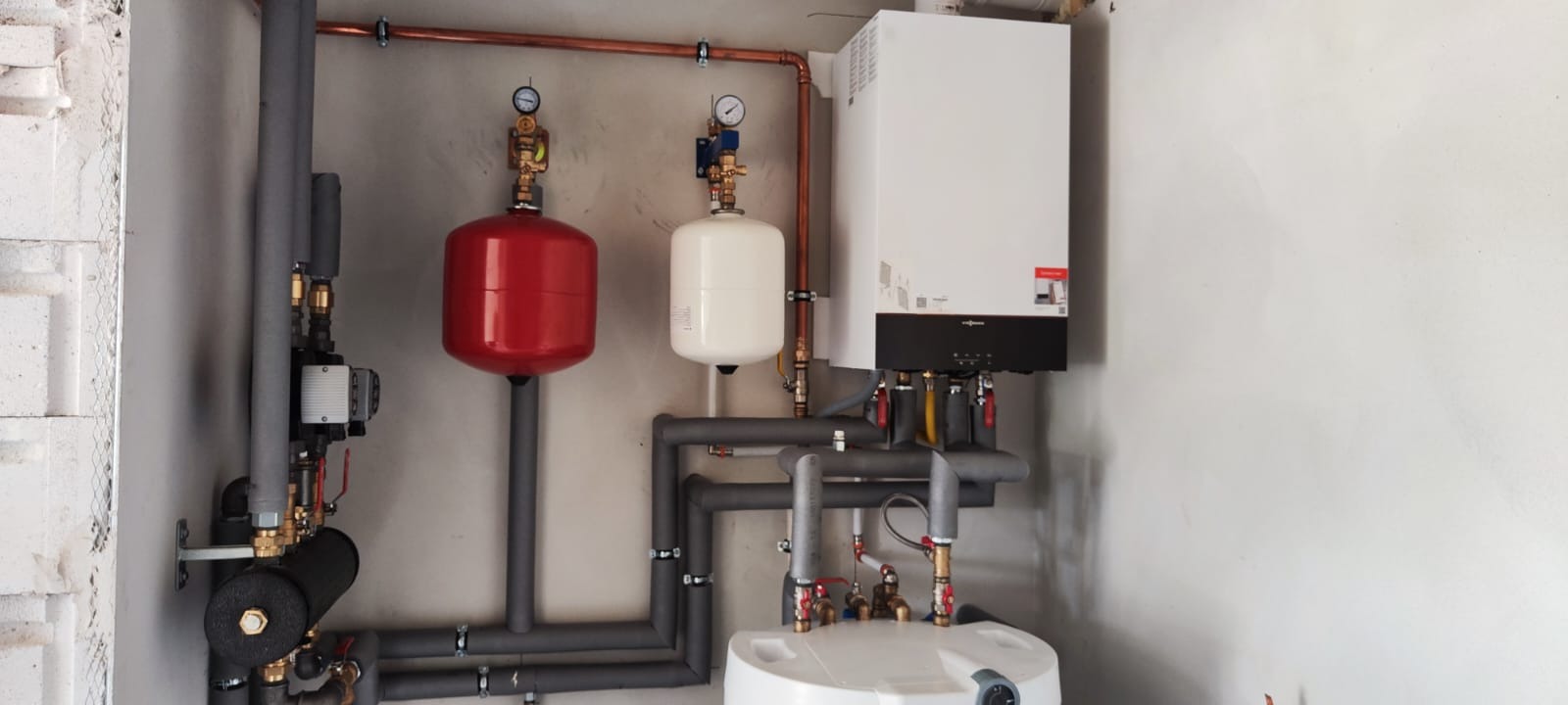 Biały piec grzewczy Viessmann z czarnym panelem, instalacja z rur w izolacji, czerwony i biały zbiornik wyrównawczy, miedziane rury na jasnej ścianie.