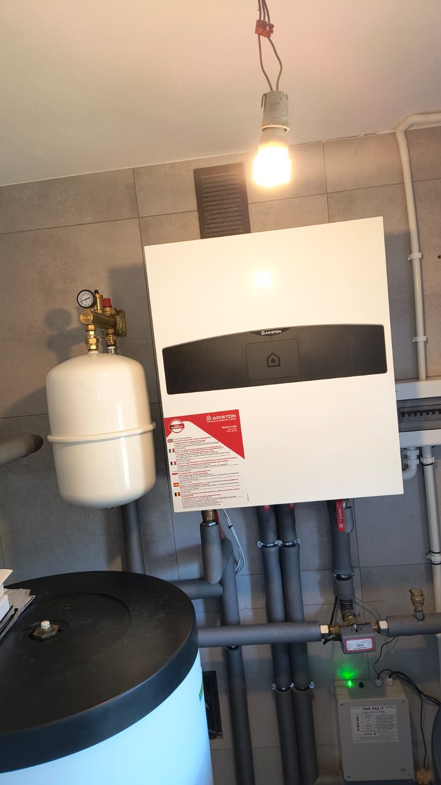 Biały, wiszący piec gazowy Ariston z podłączonymi rurami i zbiornikiem wyrównawczym, zamontowany na szarej, kafelkowej ścianie w pomieszczeniu. Widoczne elementy instalacji.
