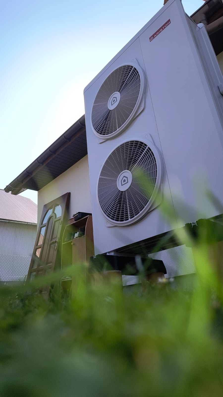 Biała pompa ciepła Ariston zamontowana przy budynku, widok z dołu, z zielenią w pierwszym planie, błękitne niebo w tle. Ujęcie z perspektywy trawy.