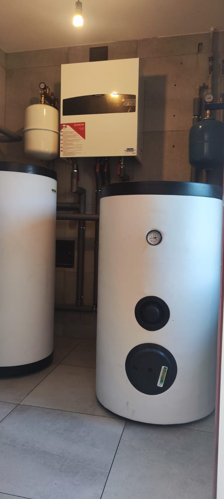 Biały piec gazowy Ariston z zbiornikami w kotłowni. Widoczne rury, zawory i manometry. Szare płytki na ścianach i podłodze. Oświetlenie górne.