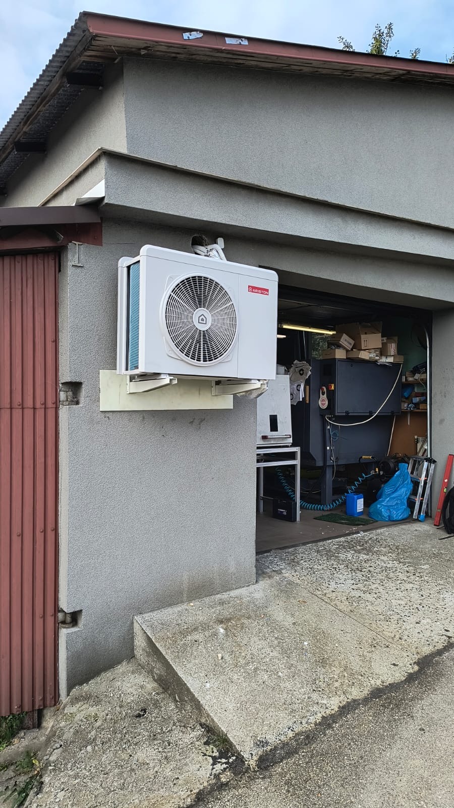 Zamontowana jednostka zewnętrzna pompy ciepła Ariston na szarej ścianie budynku z otwartym garażem w tle. Widoczny podest wejściowy i fragment drzwi.