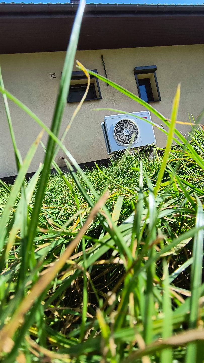 Szara pompa ciepła zamontowana na elewacji budynku, widoczna z perspektywy trawy. Dwa okna z roletami w tle. Ujęcie z dołu, blisko trawy, z niskiej perspektywy.