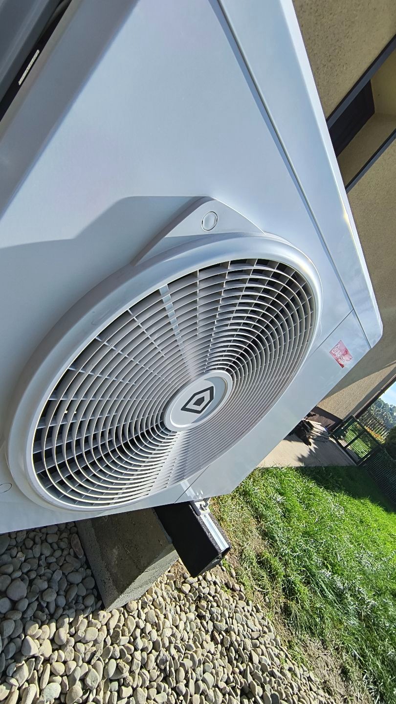 Biała pompa ciepła z widocznym wentylatorem i logo producenta, zamontowana na zewnątrz budynku, na tle trawy i kamieni.