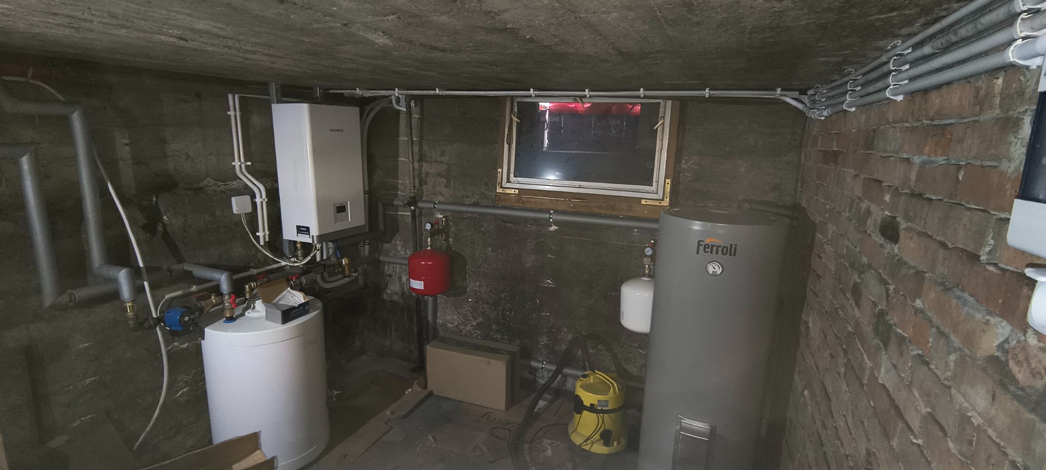 Piwnica z zamontowanym kotłem gazowym Kaisai, zasobnikiem Ferroli, instalacją hydrauliczną i oknem. Widoczne rury i akcesoria. Ściany z cegły i betonu.