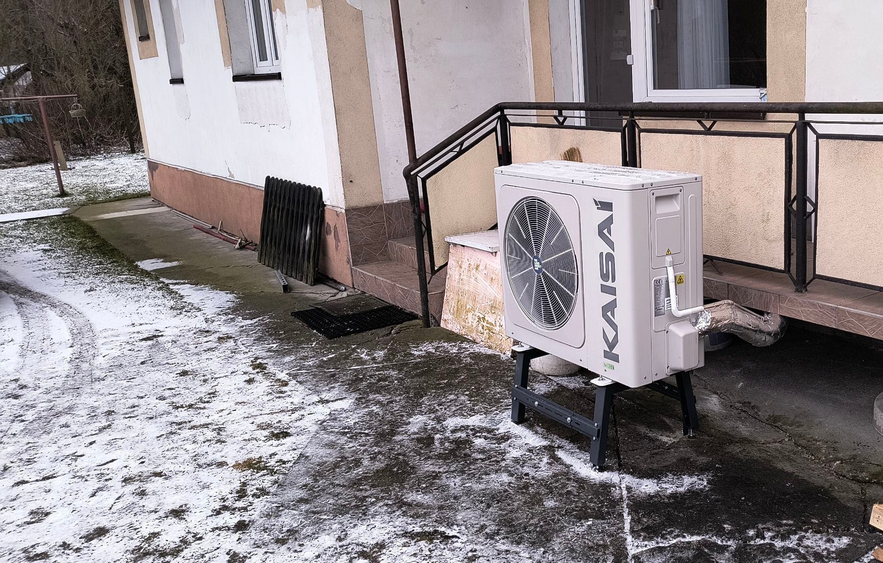 Biała pompa ciepła Kaisai na zewnątrz budynku z widocznymi rurami instalacyjnymi, ustawiona na czarnej konstrukcji. W tle fragment elewacji i schody.