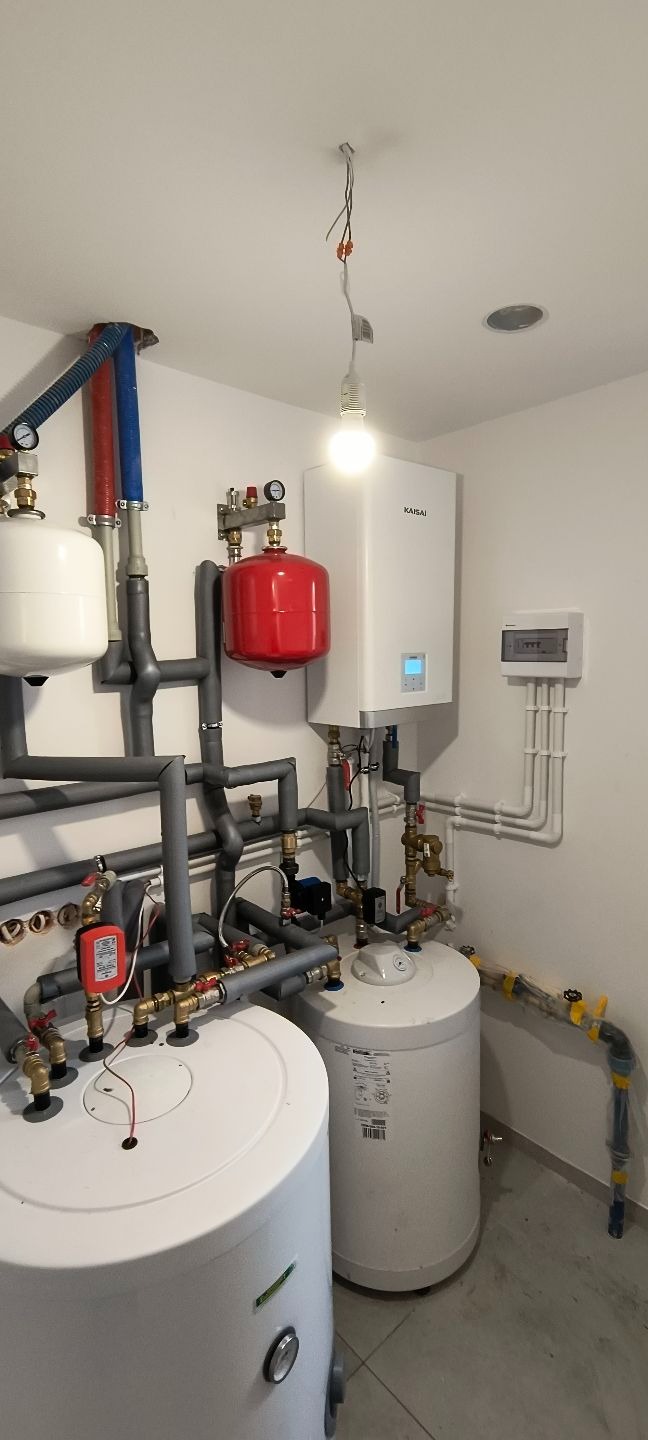 Kompletna instalacja grzewcza z kotłem Kaisai, zbiornikami, rurami i zaworami w jasnym pomieszczeniu. Widoczne elementy elektryczne i oświetlenie.