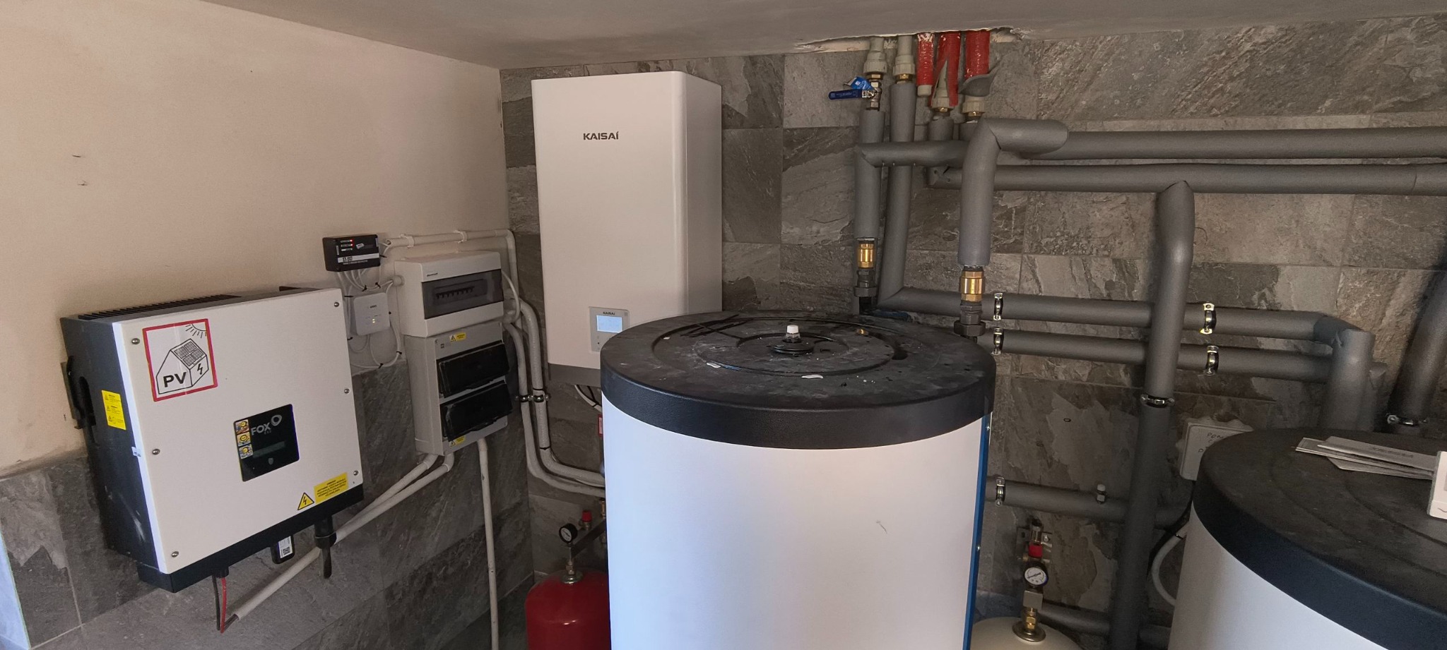 Instalacja Kaisai z zbiornikiem, inwerterem PV FoxEss i rurami w szarym oplocie na tle ściany z szarej mozaiki. Elementy instalacji grzewczej w kotłowni.