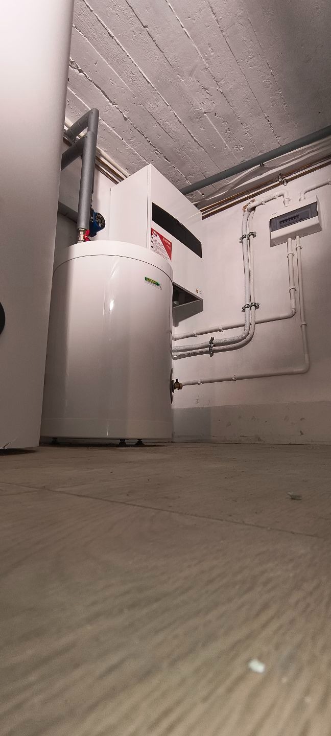 Biały zbiornik na wodę i piec w piwnicy z widocznymi rurami instalacji. Niski kąt ujęcia podkreśla surowość pomieszczenia.