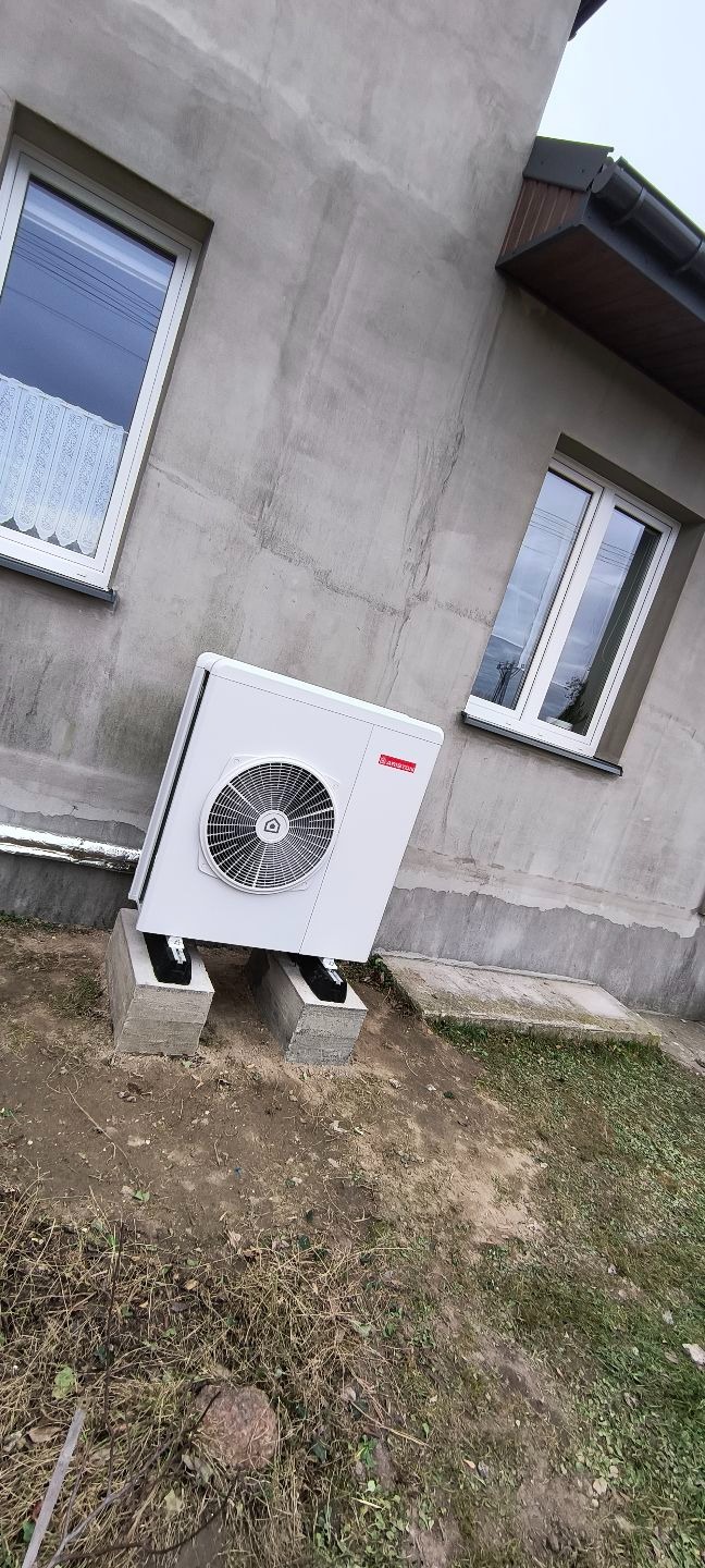 Biała pompa ciepła Ariston zamontowana na betonowych blokach przy szarej ścianie budynku z dwoma oknami. Widoczne elementy instalacji.