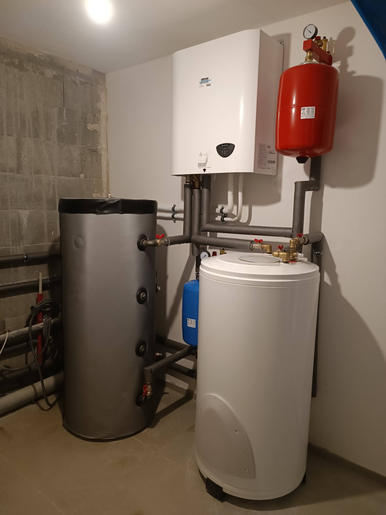Kompletna instalacja grzewcza w kotłowni: biały piec gazowy, czerwony zbiornik wyrównawczy, szary i biały zasobnik wody, rury w izolacji. Ściana surowa i pomalowana.