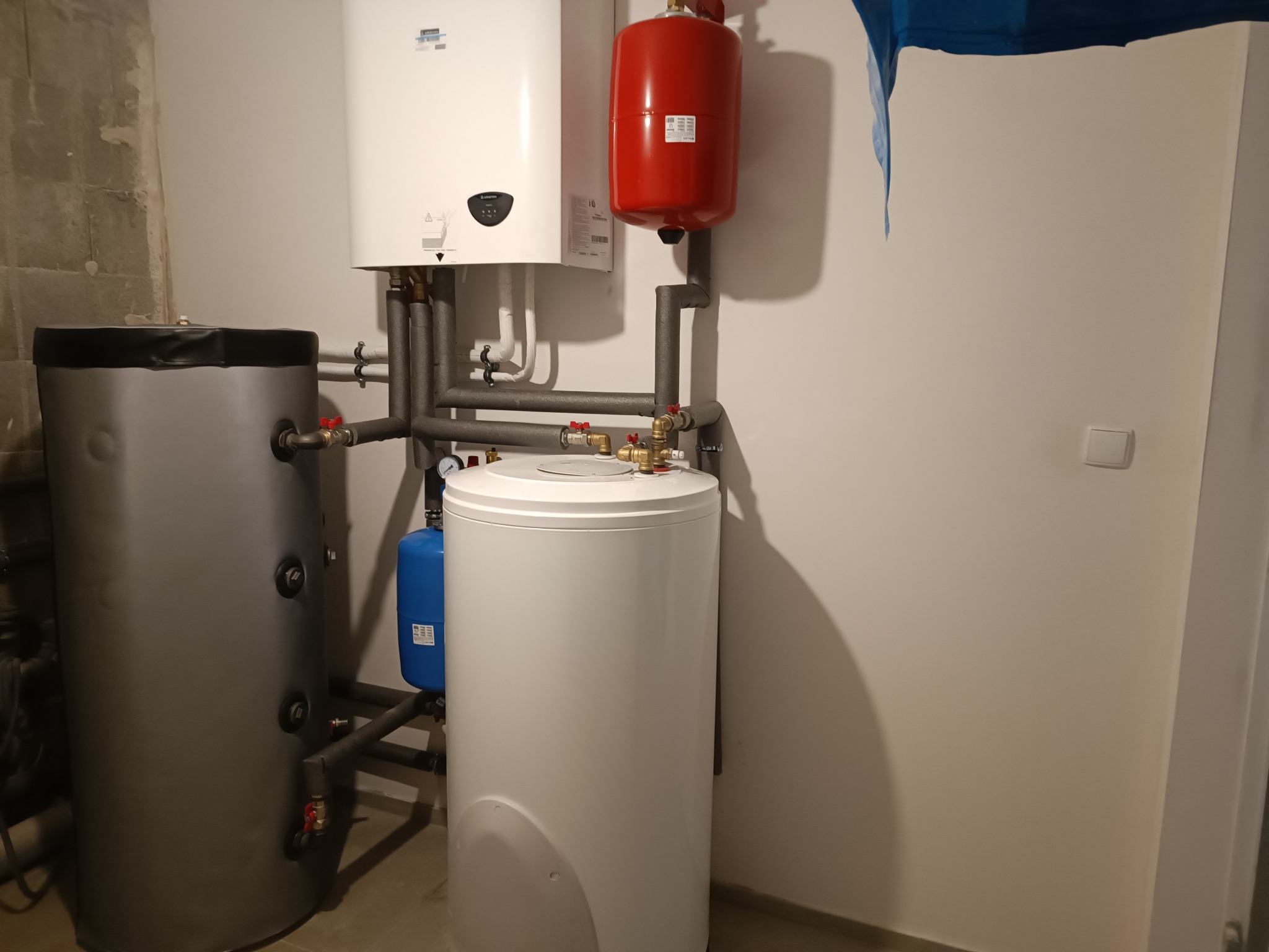 Zestaw urządzeń grzewczych: biały piec gazowy, czerwony zbiornik wyrównawczy, szary zasobnik ciepłej wody i niebieski zbiornik przeponowy. Instalacja w kotłowni.