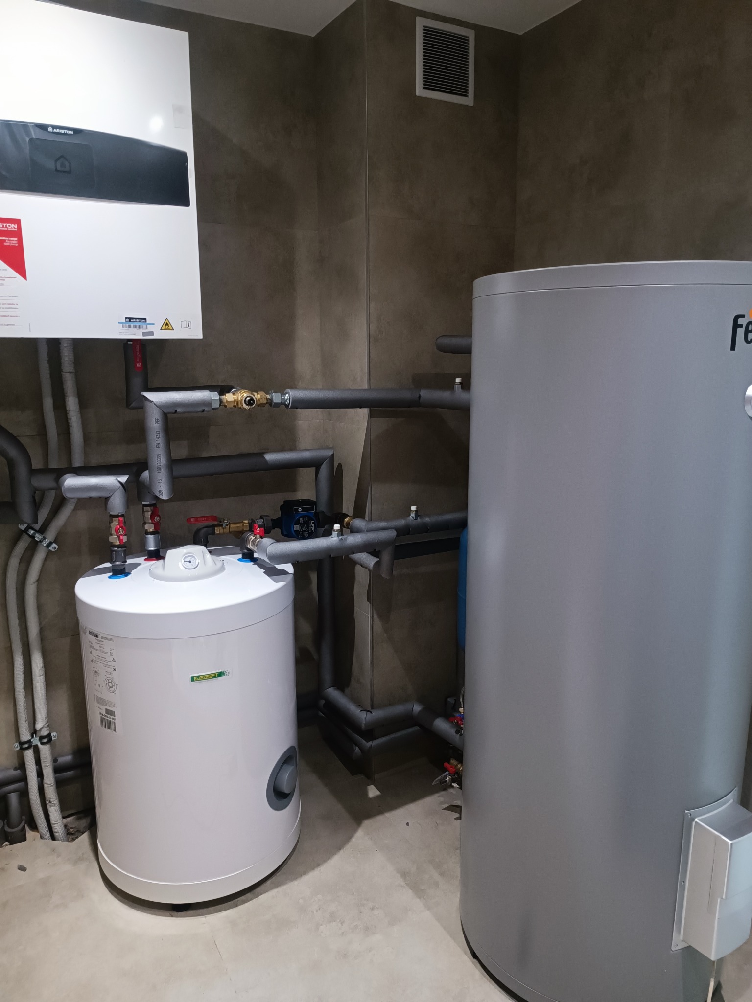 Biały zasobnik ciepłej wody użytkowej i szary zbiornik buforowy w kotłowni, z widocznymi rurami i armaturą. Na ścianie wisi piec gazowy Ariston.
