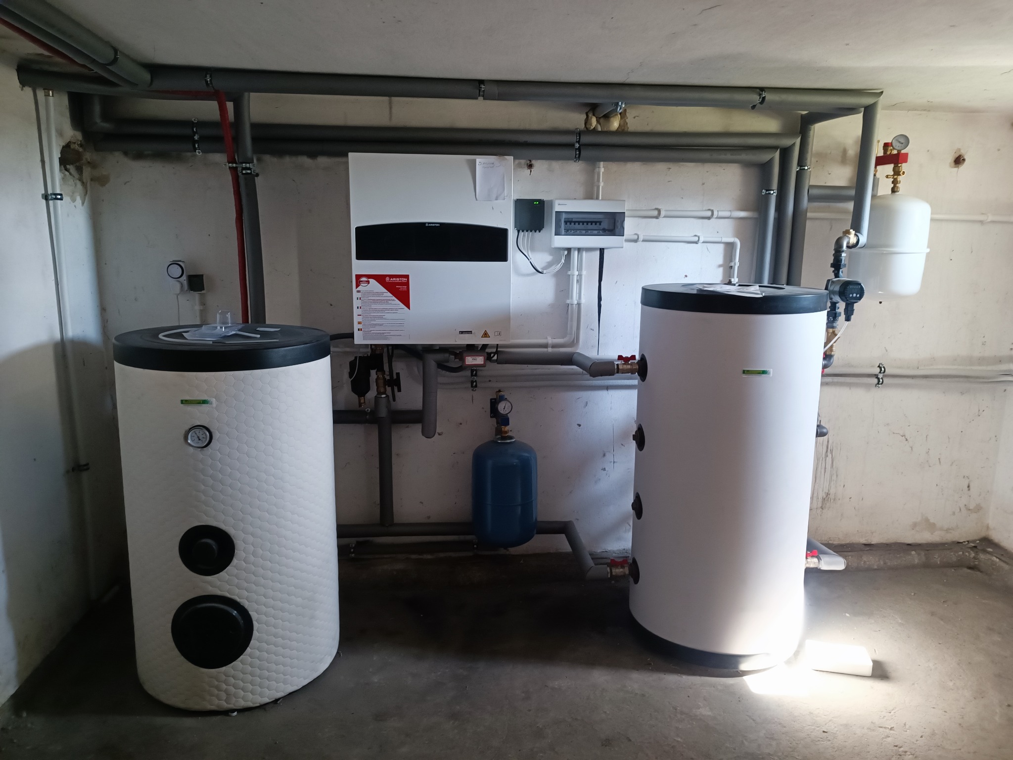 Zestaw urządzeń grzewczych w kotłowni: dwa białe zbiorniki, piec z czarnym panelem, rozdzielnica elektryczna i rury instalacji grzewczej. Widok na kompletną instalację.