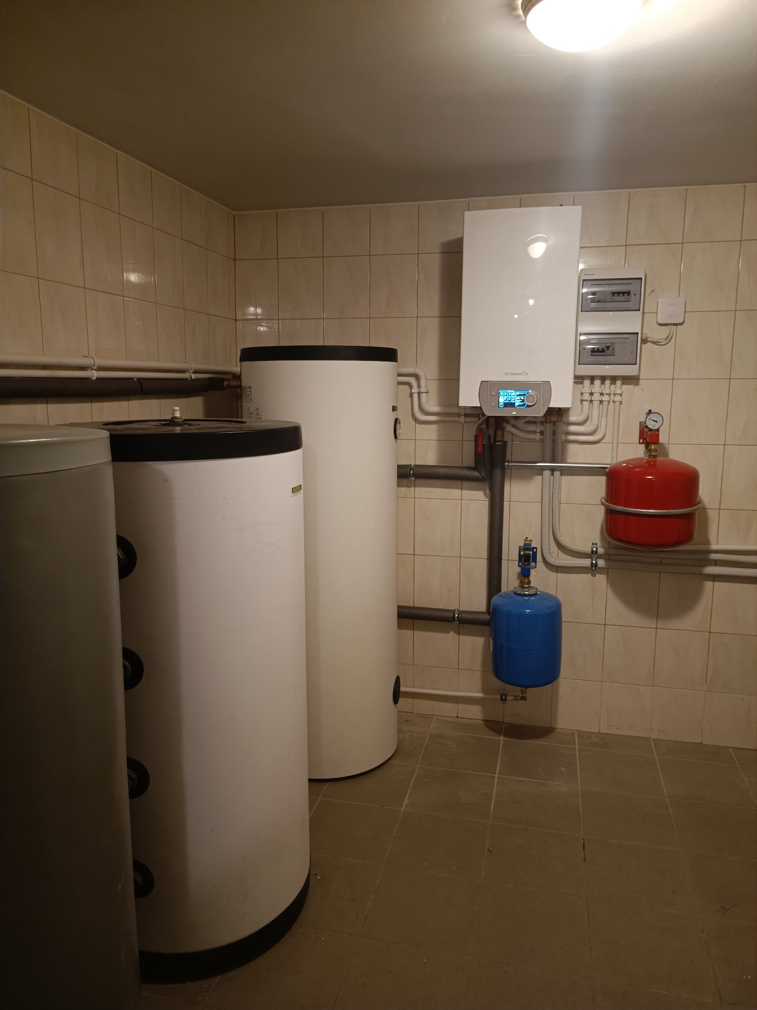Kompletna instalacja grzewcza w kotłowni: biały piec gazowy De Dietrich, dwa białe i jeden szary zbiornik akumulacyjny, czerwony i niebieski zbiornik przeponowy.