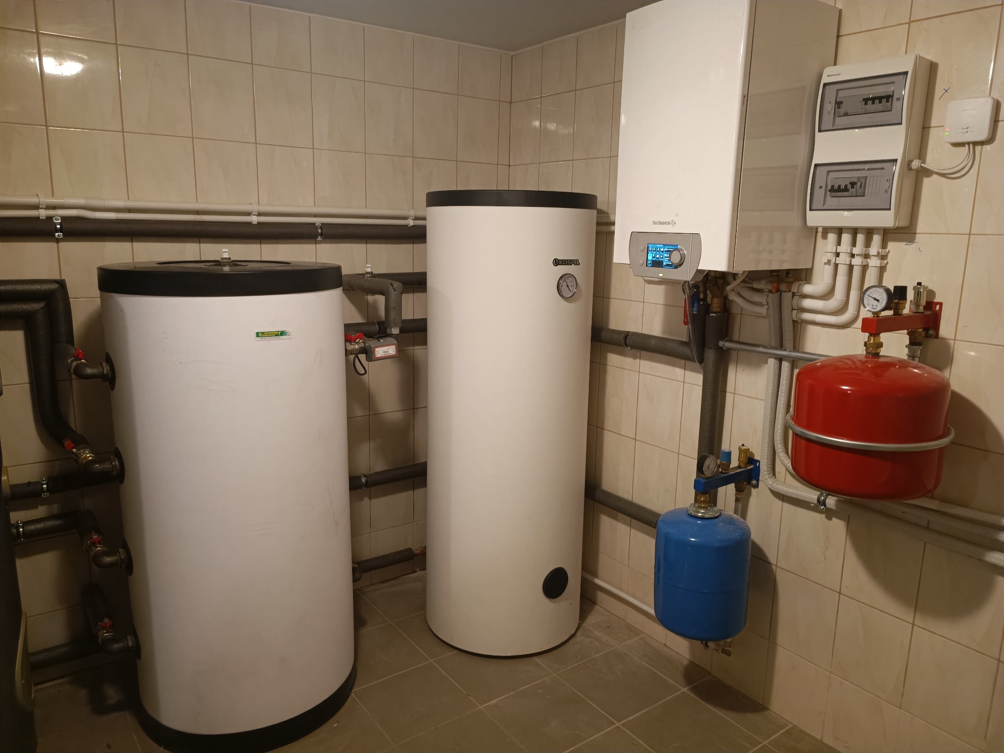 Kompletna instalacja grzewcza w kotłowni: dwa białe zbiorniki akumulacyjne, piec gazowy De Dietrich, czerwony i niebieski zbiornik wyrównawczy, rury i armatura na tle beżowych płytek.