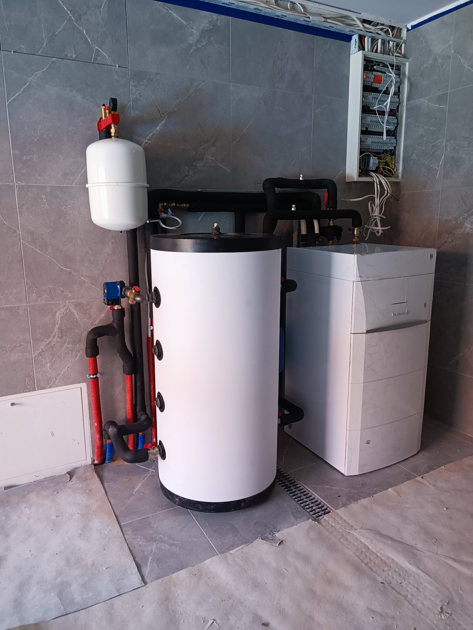 Biały zasobnik ciepłej wody użytkowej z podłączonymi rurami, obok piec gazowy w folii ochronnej, na tle szarych płytek. Widoczna skrzynka elektryczna.