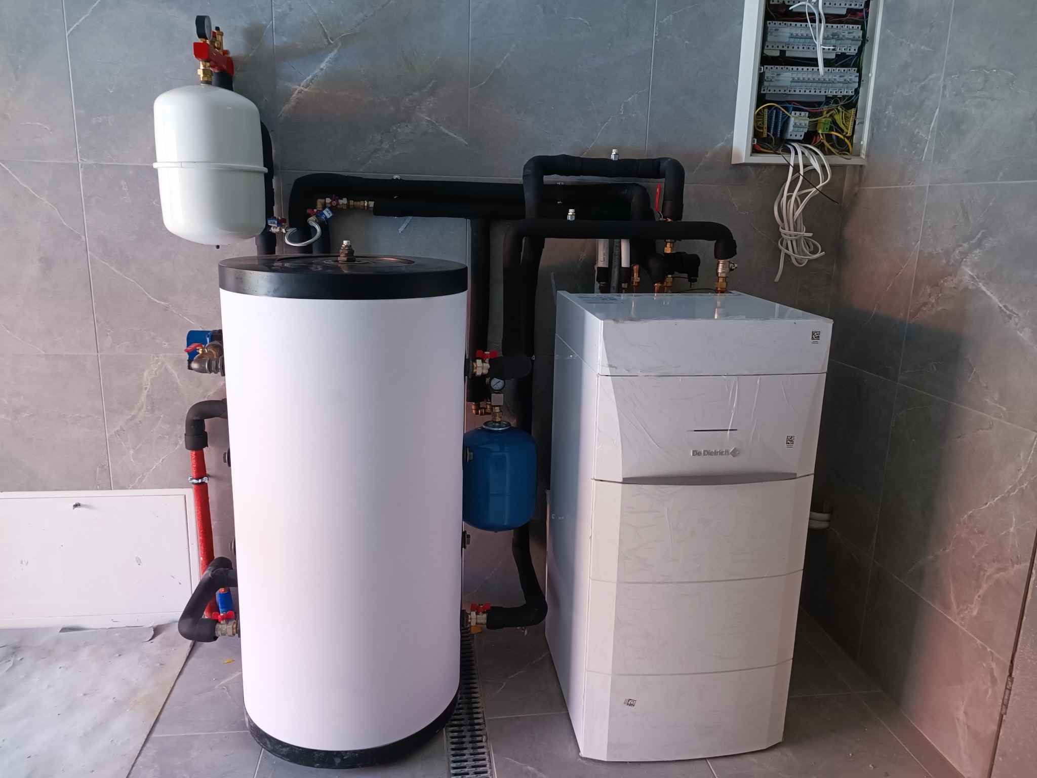 Nowoczesna kotłownia z białym zasobnikiem ciepłej wody i piecem gazowym De Dietrich, czarne rury izolacyjne, szara płytka na ścianie i podłodze, skrzynka elektryczna.