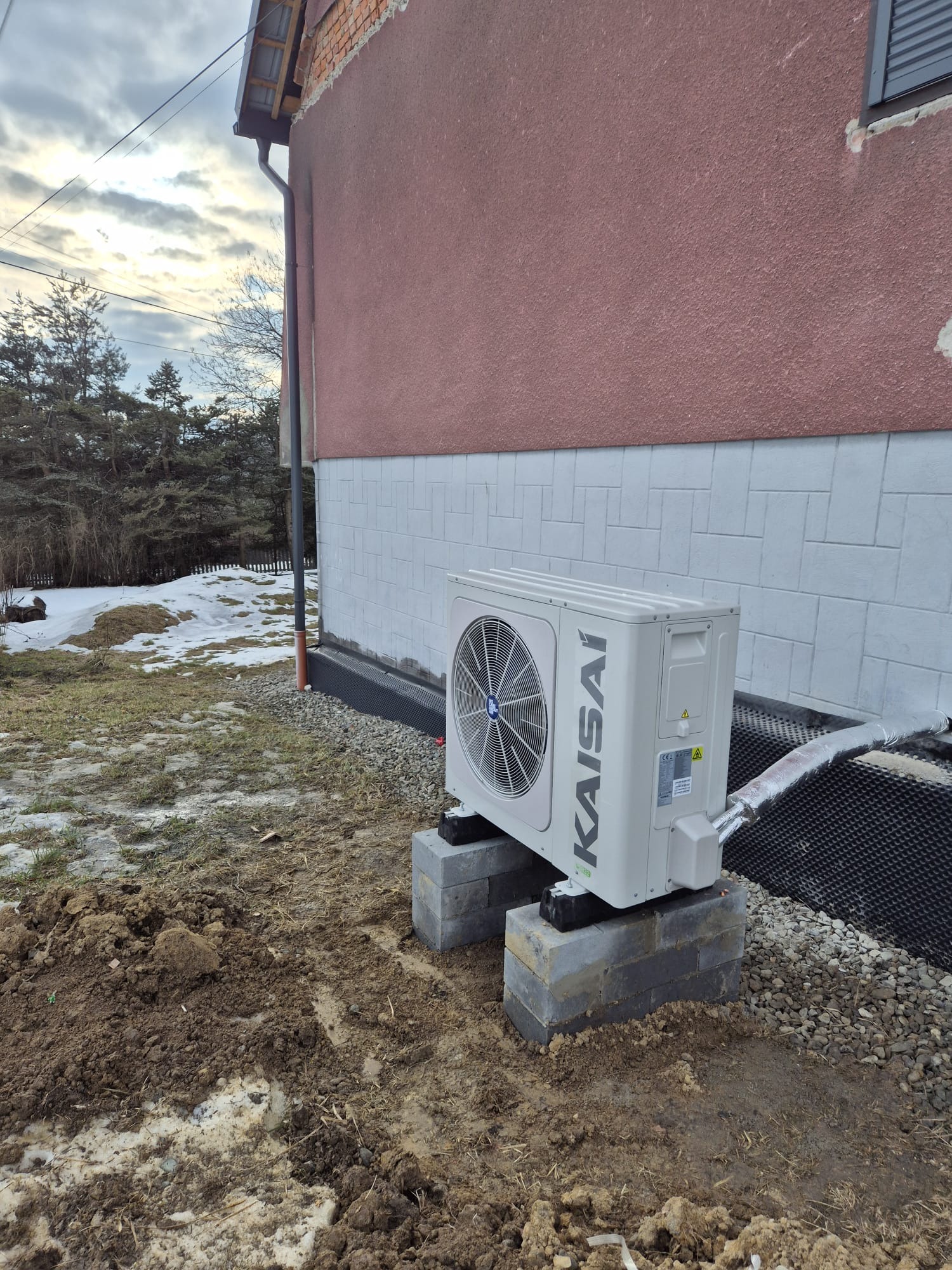 Biała pompa ciepła Kaisai zamontowana na betonowych blokach przy ścianie budynku z widoczną izolacją. W tle resztki śniegu i drzewa.