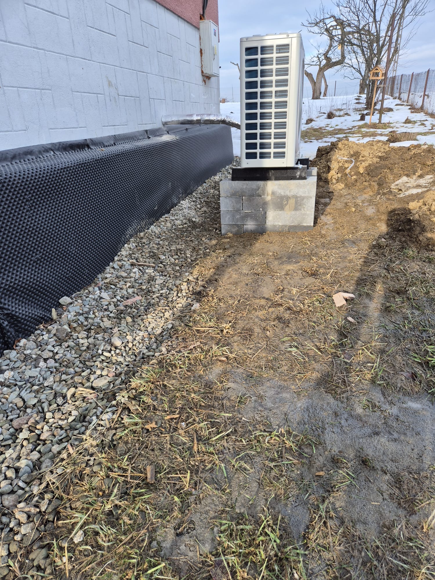 Zewnętrzna jednostka pompy ciepła na betonowej podstawie, obok ściany budynku z izolacją przeciwwilgociową i drenażem z kamieni. Widoczny fragment ogrodu.