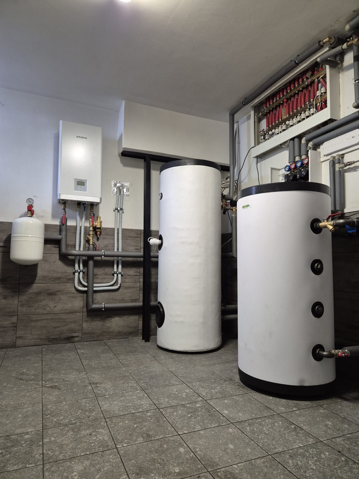 Biały kocioł Kaisai, dwa zbiorniki akumulacyjne z czarnymi podstawami i rozdzielacz instalacji grzewczej w kotłowni z szarą podłogą z płytek.