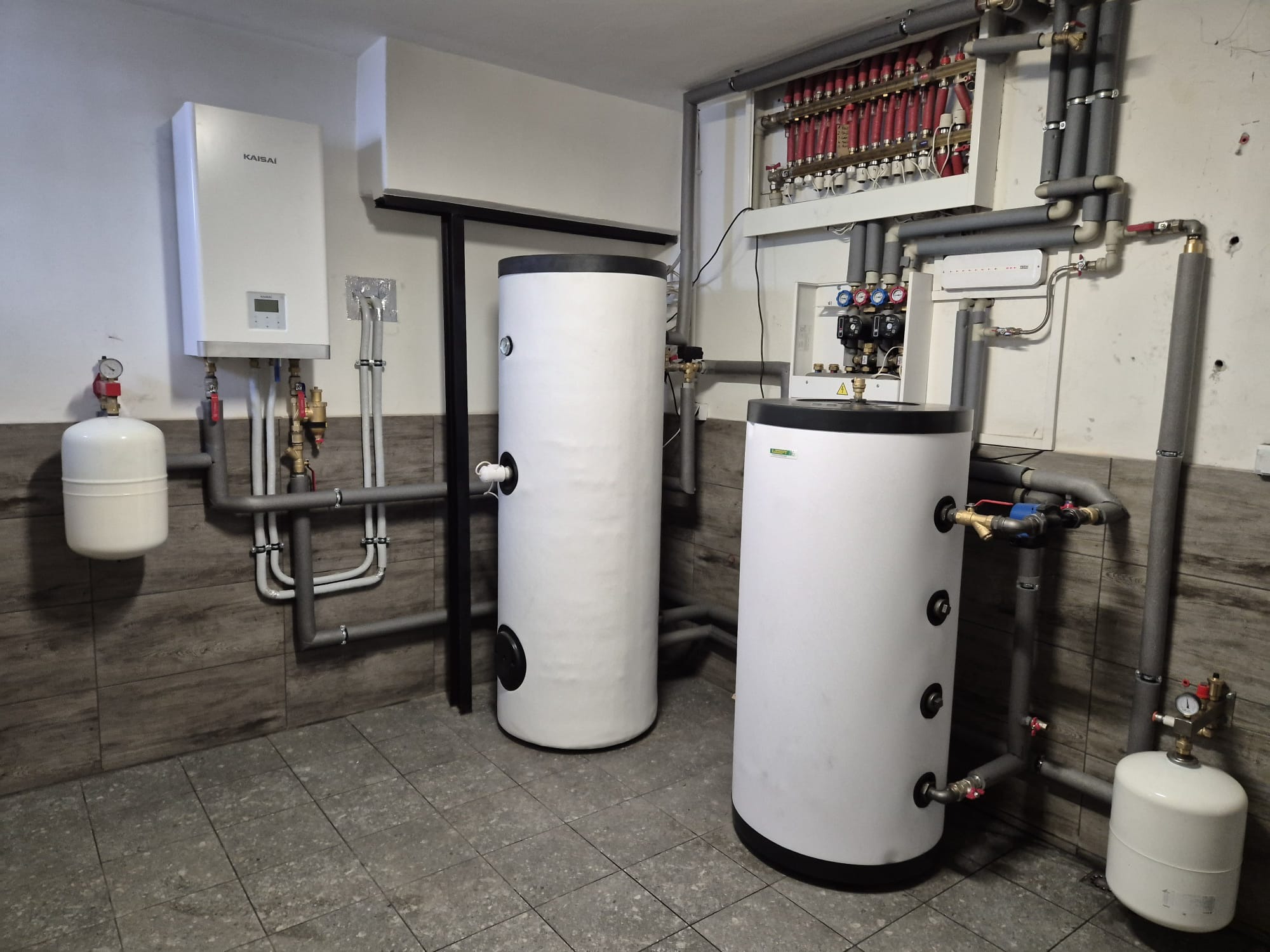 Kompletna instalacja grzewcza w kotłowni: biały piec Kaisai, dwa białe zbiorniki akumulacyjne, rozdzielacz z czerwonymi zaworami i szare rury z izolacją.