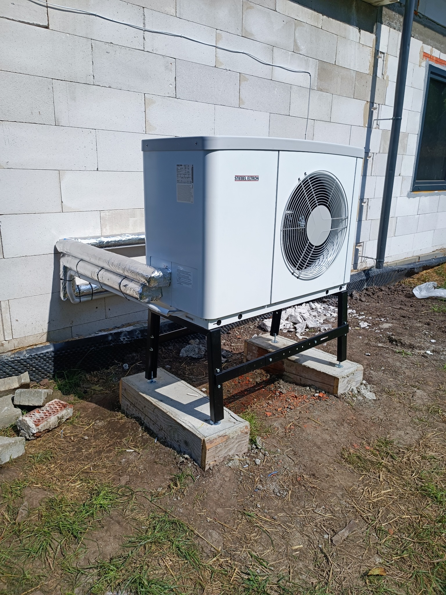 Biała pompa ciepła Stiebel Eltron na czarnym stelażu, zamontowana przy ścianie z białego bloczka. Widoczne ocieplone rury i betonowe podstawy. Teren wokół w trakcie prac.