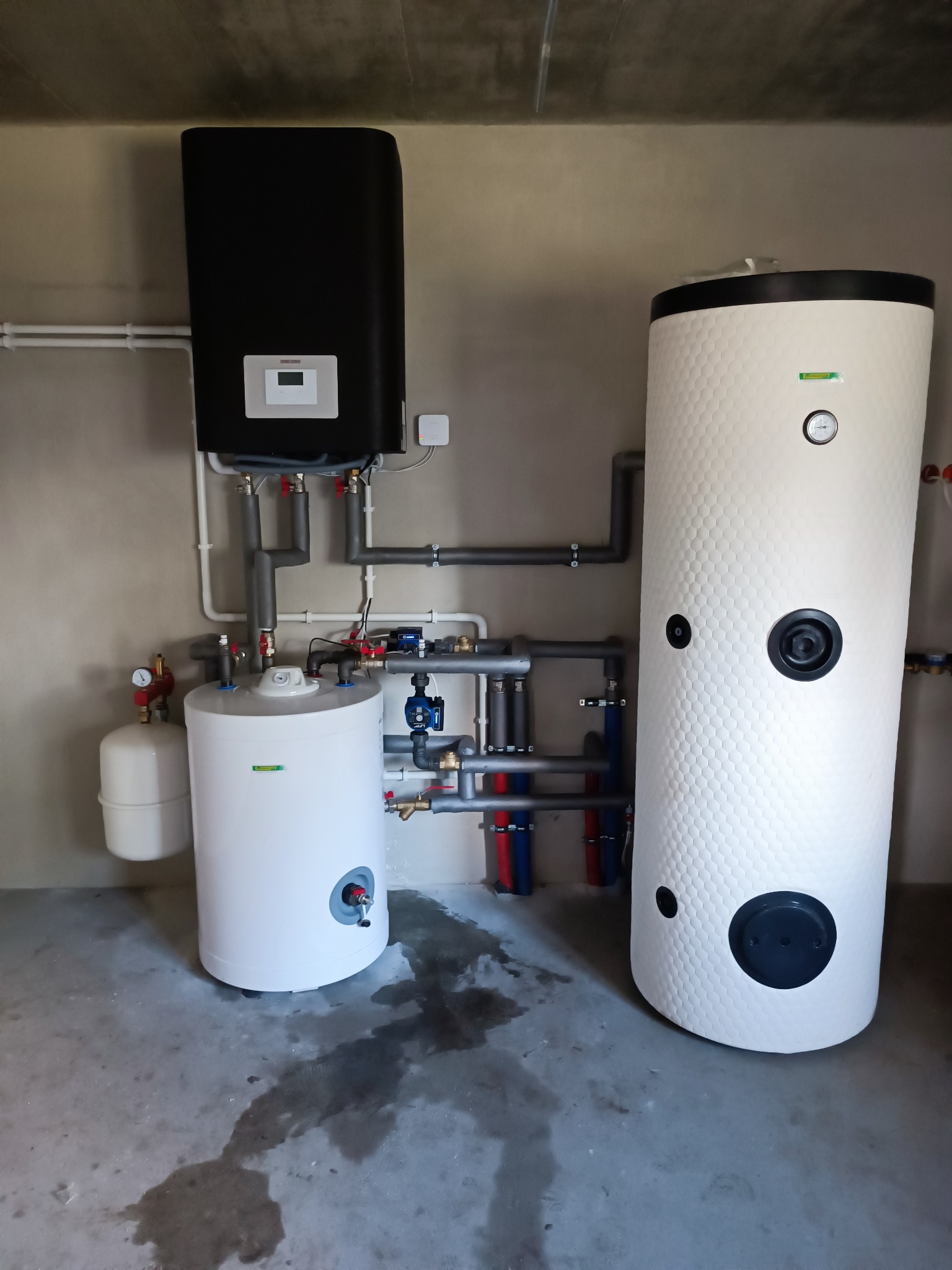 Zestaw urządzeń grzewczych: czarny zasobnik, biały zbiornik z fakturą plastra miodu, biały zbiornik z osprzętem, połączone rurami w szarej kotłowni.