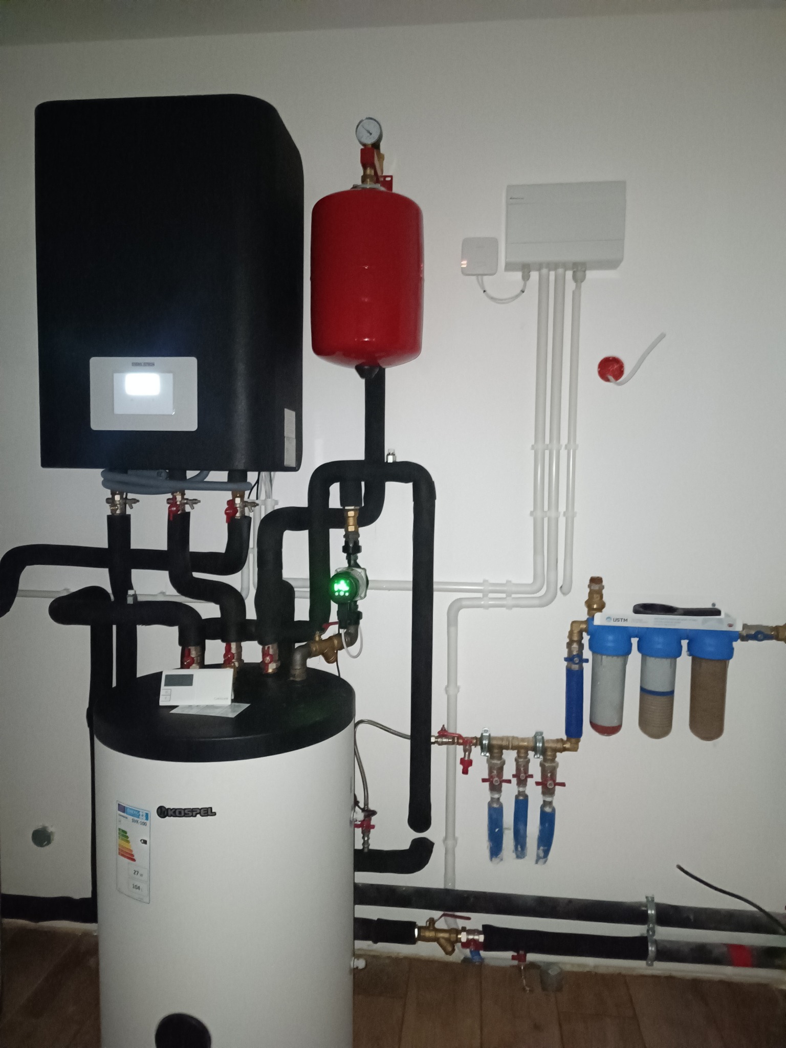 Nowoczesna instalacja grzewcza z czarnym piecem i białym zasobnikiem Kospel, połączona rurami. Widoczne filtry wody i elementy sterujące na białej ścianie.