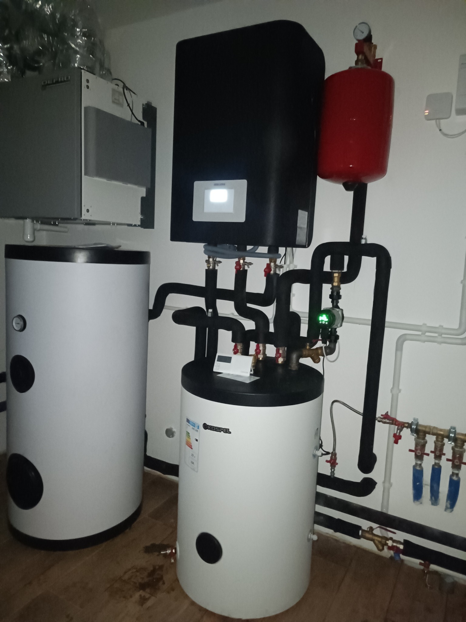 Zestaw urządzeń grzewczych: czarny piec z wyświetlaczem, czerwony zbiornik wyrównawczy, białe zasobniki wody. Rury, zawory i manometr. Widok z niskiego kąta.