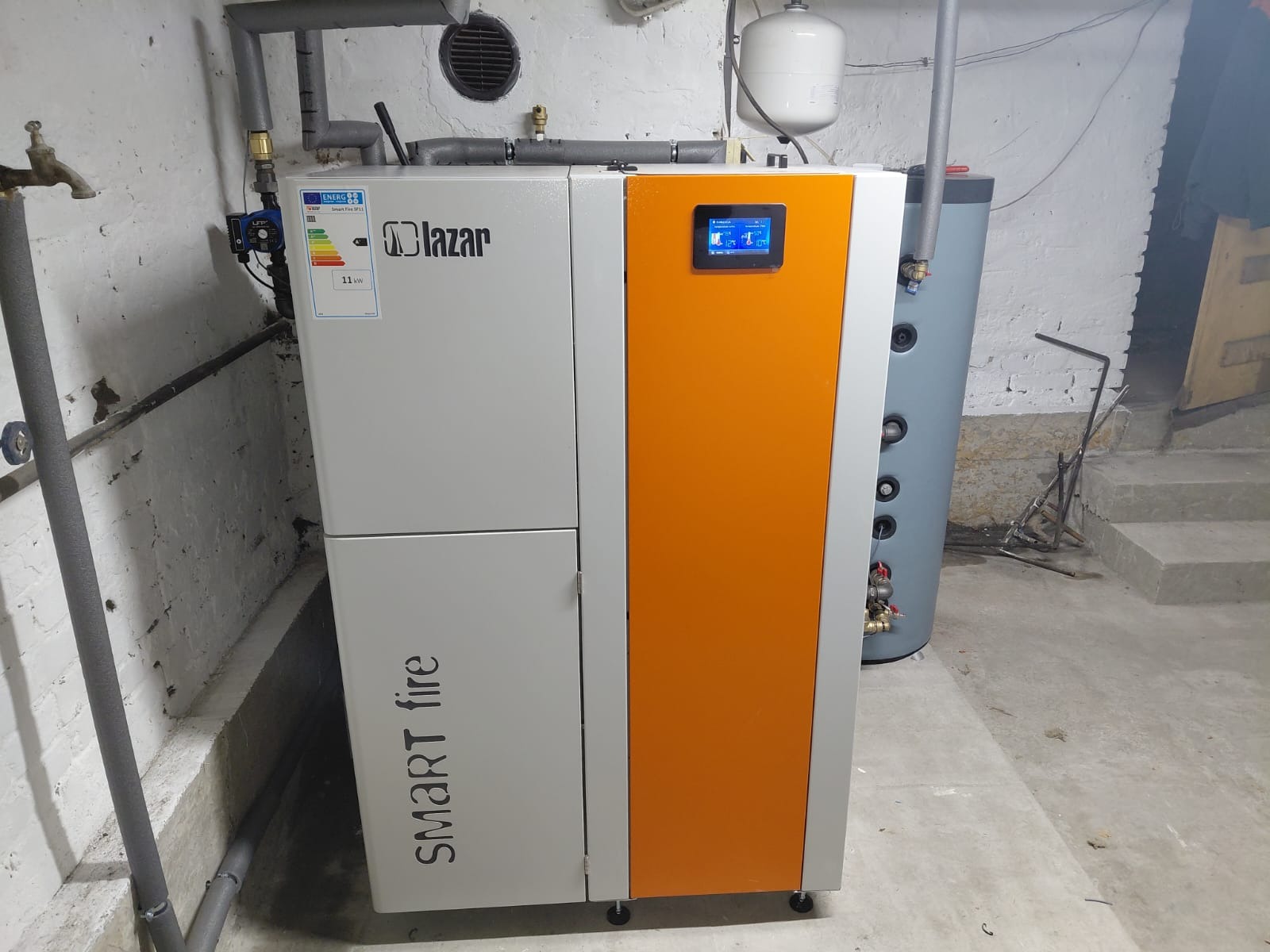 Kocioł na paliwo stałe Lazzar Smart Fire z pomarańczowym panelem, zainstalowany w piwnicy. Widoczne rury, zbiornik buforowy i schody.