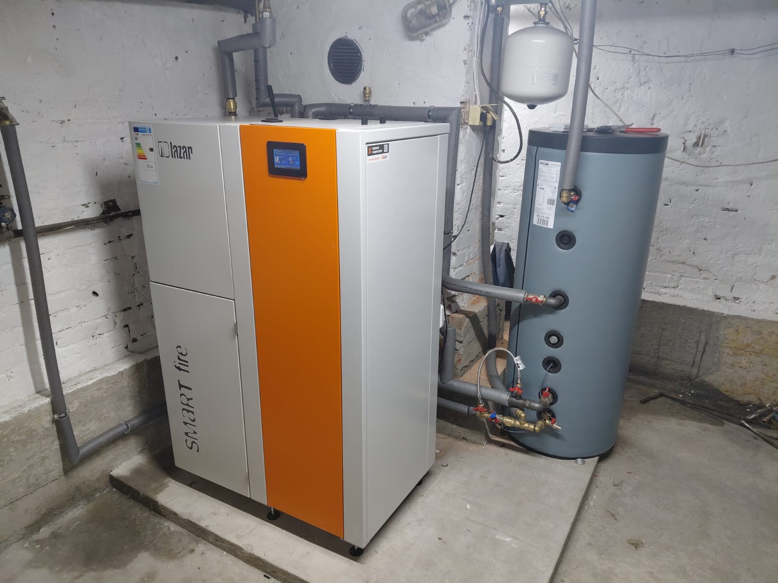 Nowoczesny, biało-pomarańczowy piec Lazar Smart Fire z zbiornikiem akumulacyjnym w kotłowni. Widoczne rury i instalacja. Surowe, białe ściany i betonowa podłoga.