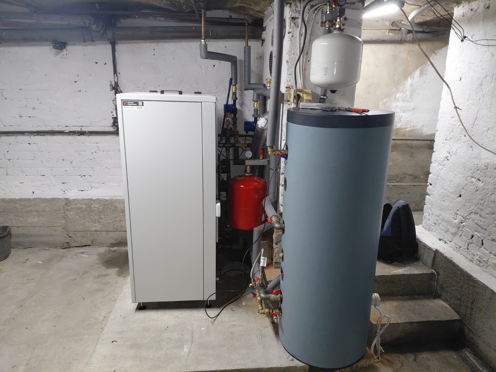 Nowoczesna instalacja grzewcza w piwnicy: biały piec, szary zasobnik wody, czerwony zbiornik wyrównawczy, rury i zawory. Widoczne schody i surowe ściany.