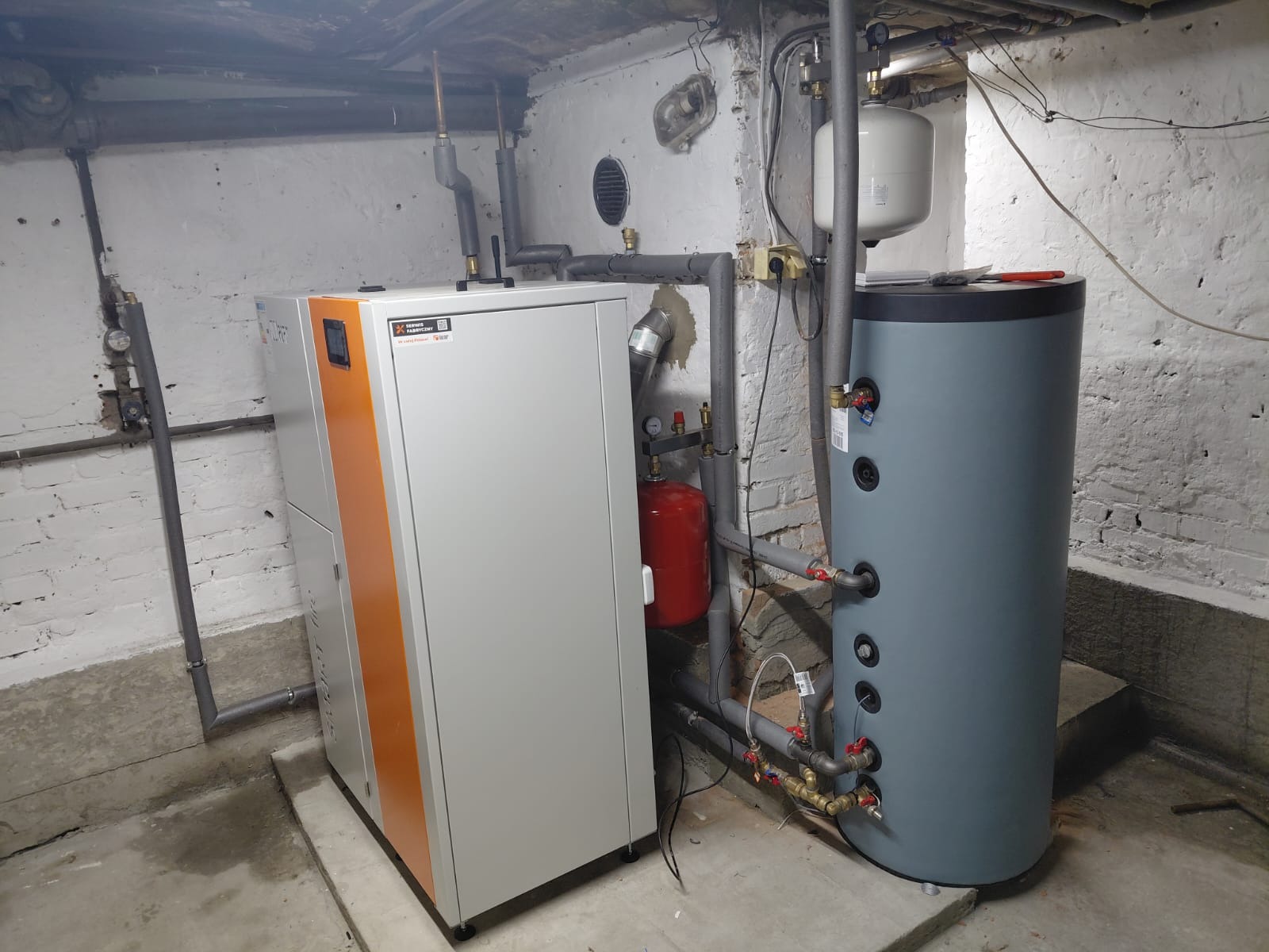 Zestaw grzewczy w piwnicy: biało-pomarańczowy piec z zasobnikiem wody i instalacją rurową. Widoczne elementy montażowe i podłączenia. Ściany z cegły.