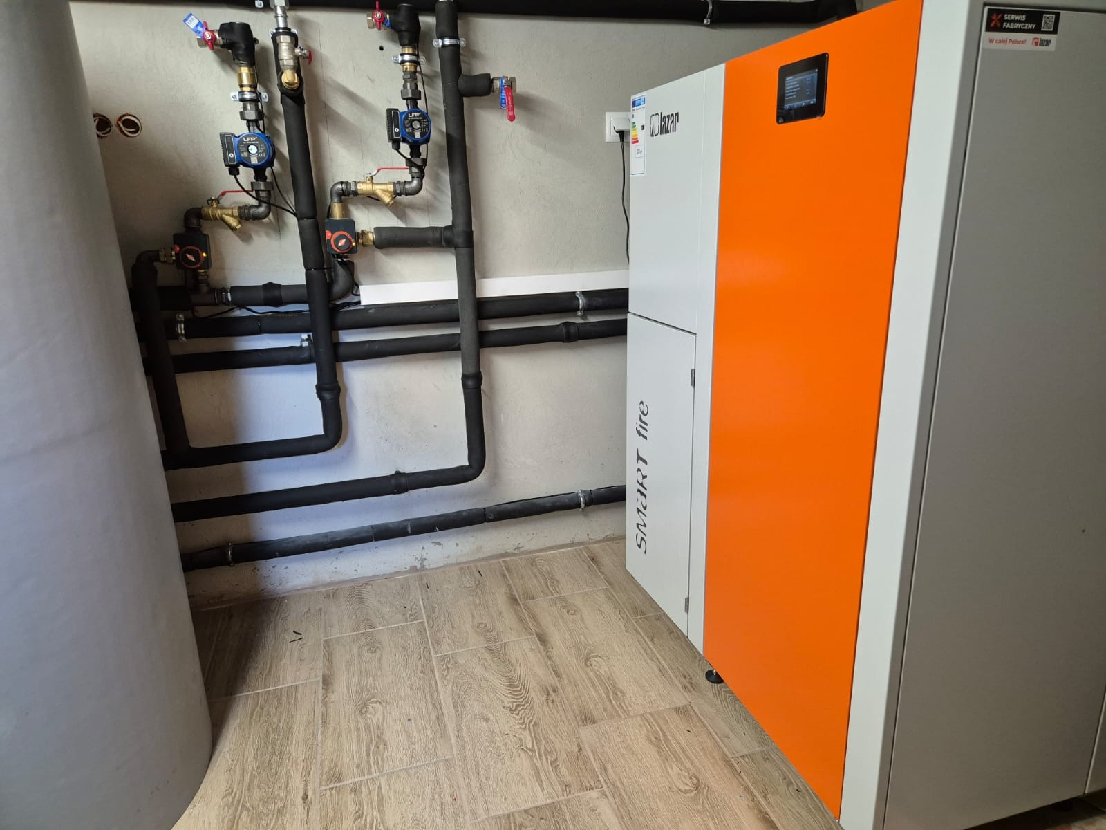 Nowoczesny, pomarańczowo-biały piec z czarnymi rurami instalacji grzewczej na tle jasnej ściany i podłogi imitującej drewno. Widoczne zawory i pompy obiegowe.