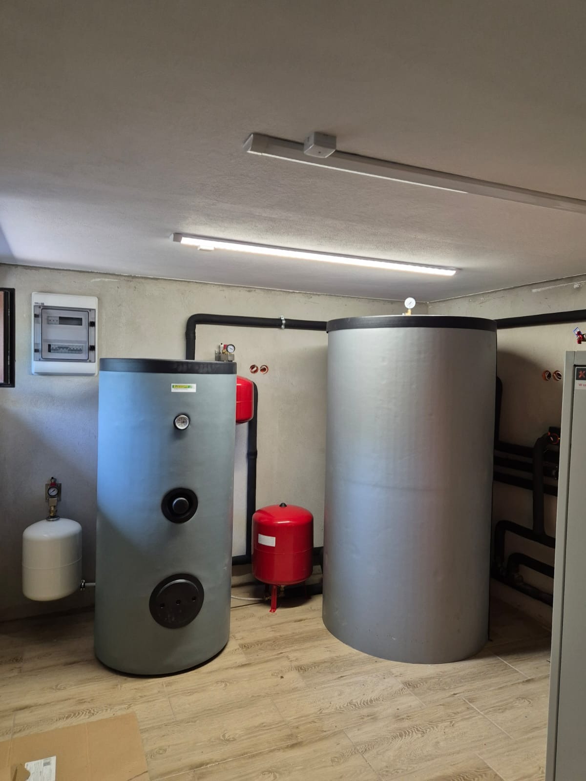 Dwa zbiorniki akumulacyjne w kotłowni, jeden szary z manometrem i czarnymi otworami rewizyjnymi, drugi srebrny, rury instalacji grzewczej na ścianie.