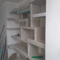 Szkielet regału z płyt kartonowo-gipsowych w trakcie montażu, widoczne profile, narzędzia i resztki materiałów na podłodze.