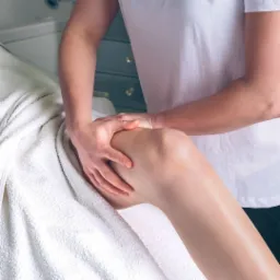 Terapeuta manualny uciska kolano pacjenta leżącego na białym ręczniku, w tle jasnoniebieska komoda z kryształowymi gałkami.