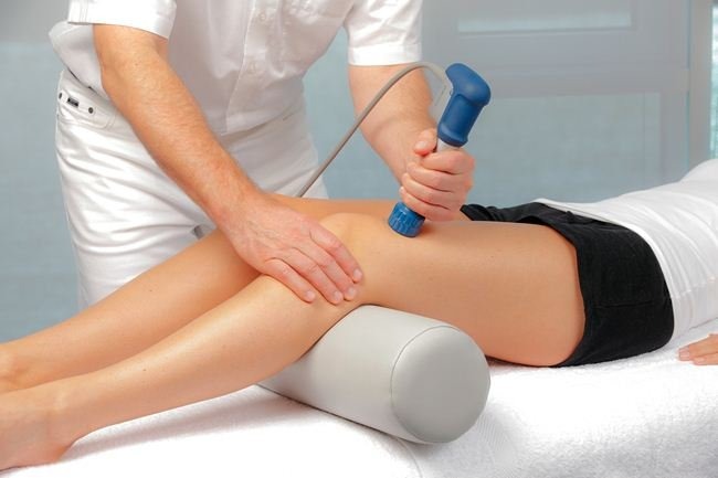 Pacjentka leżąca na leżance, pod kolanem wałek, terapeuta wykonuje zabieg z użyciem niebieskiego urządzenia z przewodem.