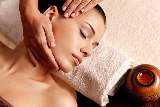 Kobieta z zamkniętymi oczami podczas masażu twarzy, z ceramicznym świecznikiem obok głowy i ręcznikiem pod głową.