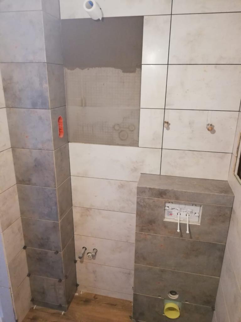 Świeżo wykafelkowana łazienka w trakcie prac wykończeniowych, widoczne odstające płytki ceramiczne, instalacja podtynkowa WC i fragment drewnopodobnej podłogi.