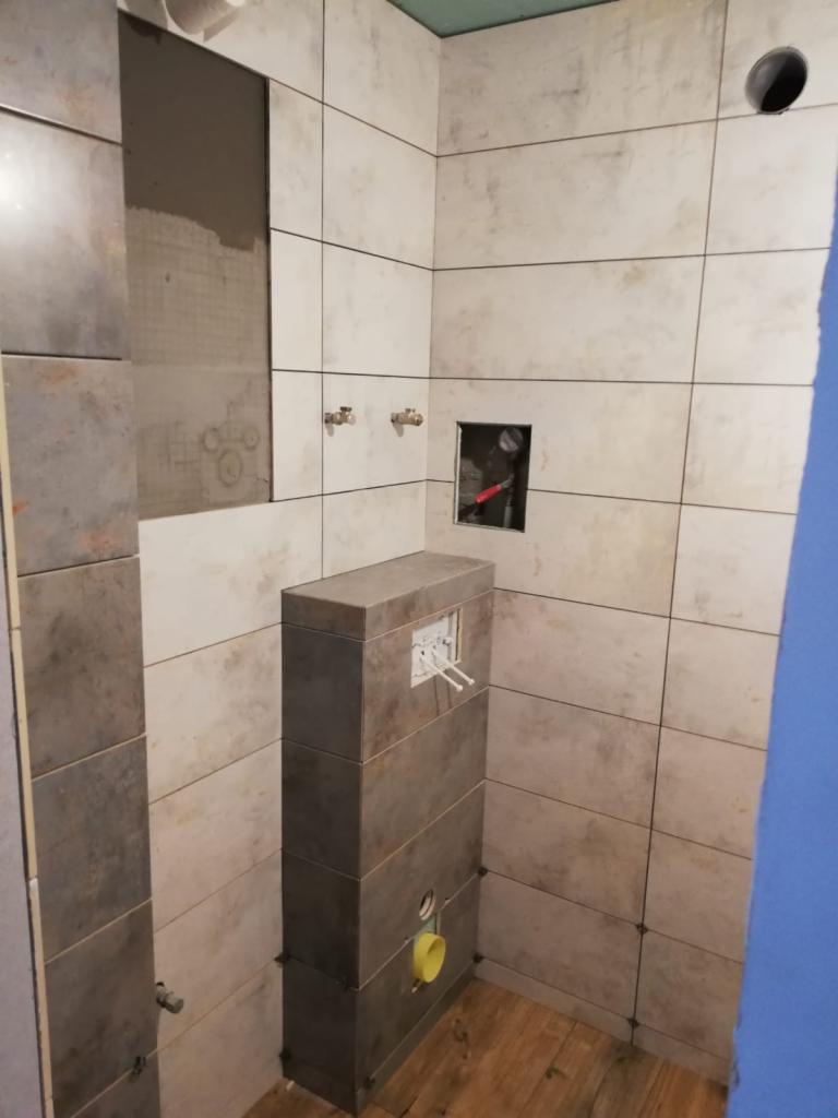 Niedokończona łazienka z częściowo położonymi kremowo-szarymi płytkami, widoczną instalacją podtynkową i fragmentem nieotynkowanej ściany.