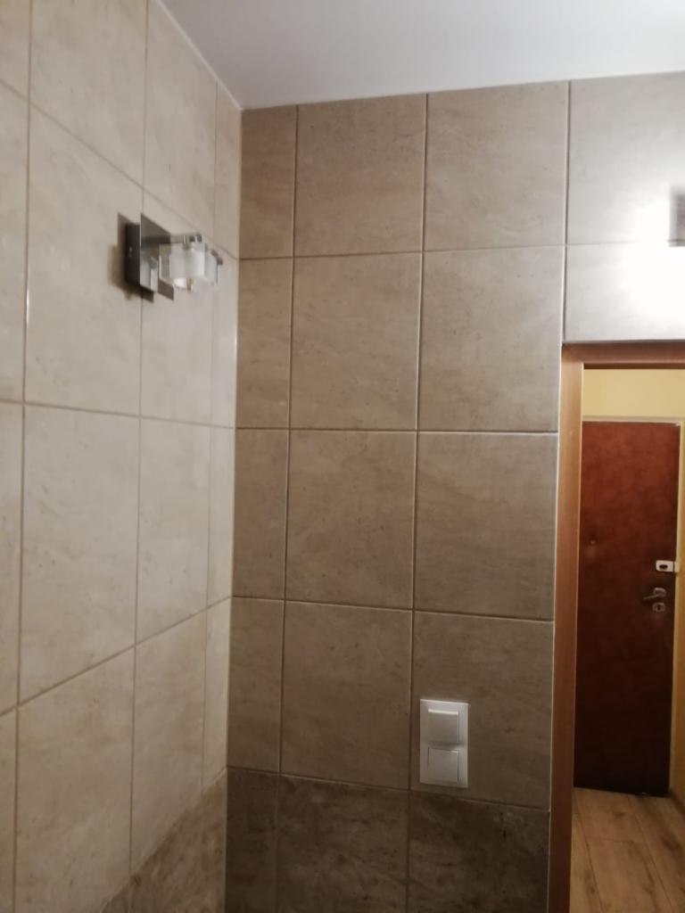 Beżowe, kwadratowe płytki ceramiczne na ścianie łazienki z chromowanym kinkietem i białym włącznikiem światła, widoczny fragment ościeżnicy i brązowe drzwi.