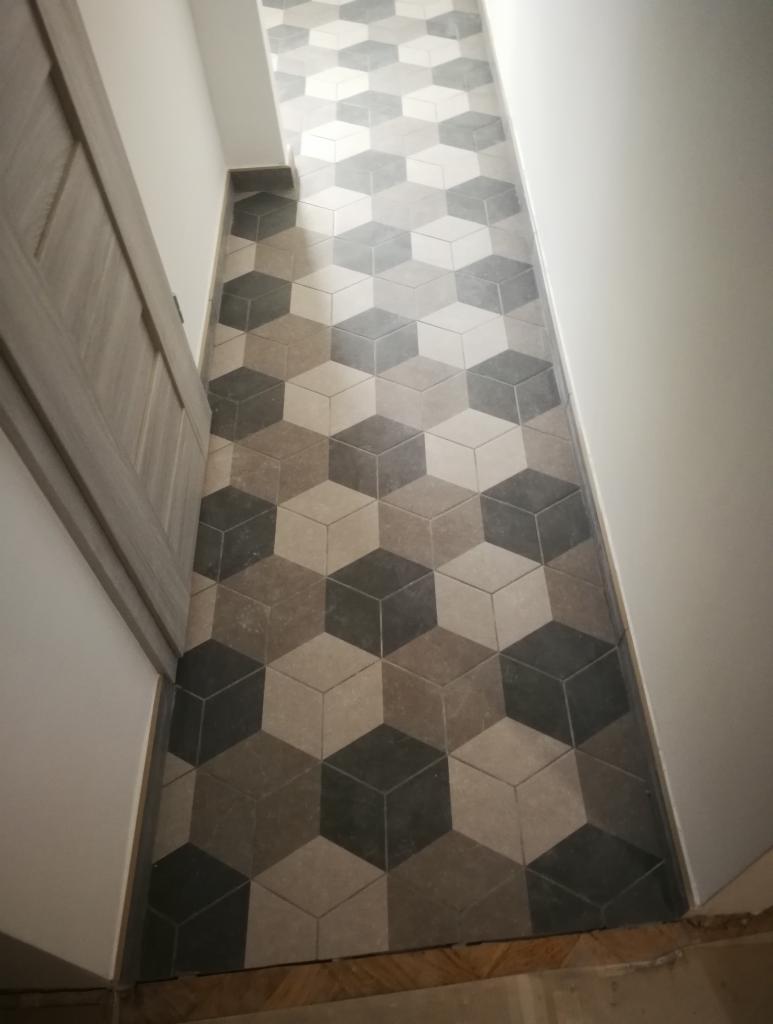 Ułożona podłoga z płytek heksagonalnych w odcieniach szarości i beżu, tworzących iluzję trójwymiarowych kostek, widoczna przy białej ścianie i drewnianej futrynie drzwi.