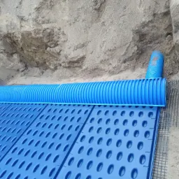 Instalacja podziemna: niebieska rura karbowana ułożona na niebieskich panelach z otworami, widoczna siatka zabezpieczająca i wykop z ziemią w tle.