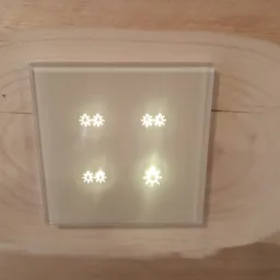 Szklany panel sterowania z podświetlanymi ikonami w kształcie płatków śniegu, zamontowany na drewnianej ścianie o widocznej strukturze słojów.